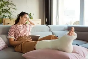 young girl sitting on her sofa at home has cast on her leg, and she looks upset/bored.