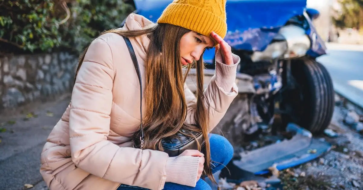 Young female driver wearing winter coat and hat crouched on ground in front of a vehicle with frontal crash damage