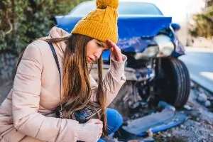 Young female driver wearing winter coat and hat crouched on ground in front of a vehicle with frontal crash damage